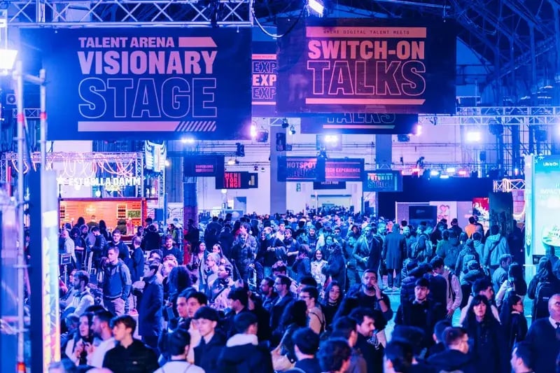 Crowded expo hall with people gathered under "Visionary Stage" and "Switch-On Talks" banners, illuminated by vibrant blue and purple lights.