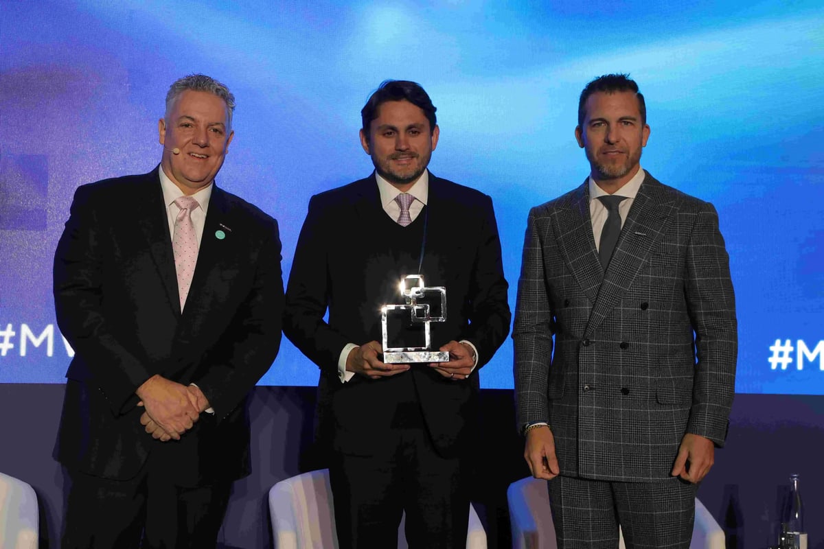 Three men in formal business attire stand on stage in front of a blue digital backdrop with the text '#MWC25MP.' The man in the center holds a shiny, geometric-shaped award, signifying an achievement. The individuals on either side smile and pose for the photo. The setting appears to be an award ceremony recognizing government leadership in 2025.